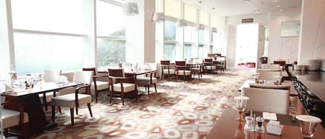 Un comedor de hotel con varias mesas y sillas, ventanas amplias y una alfombra estampada.