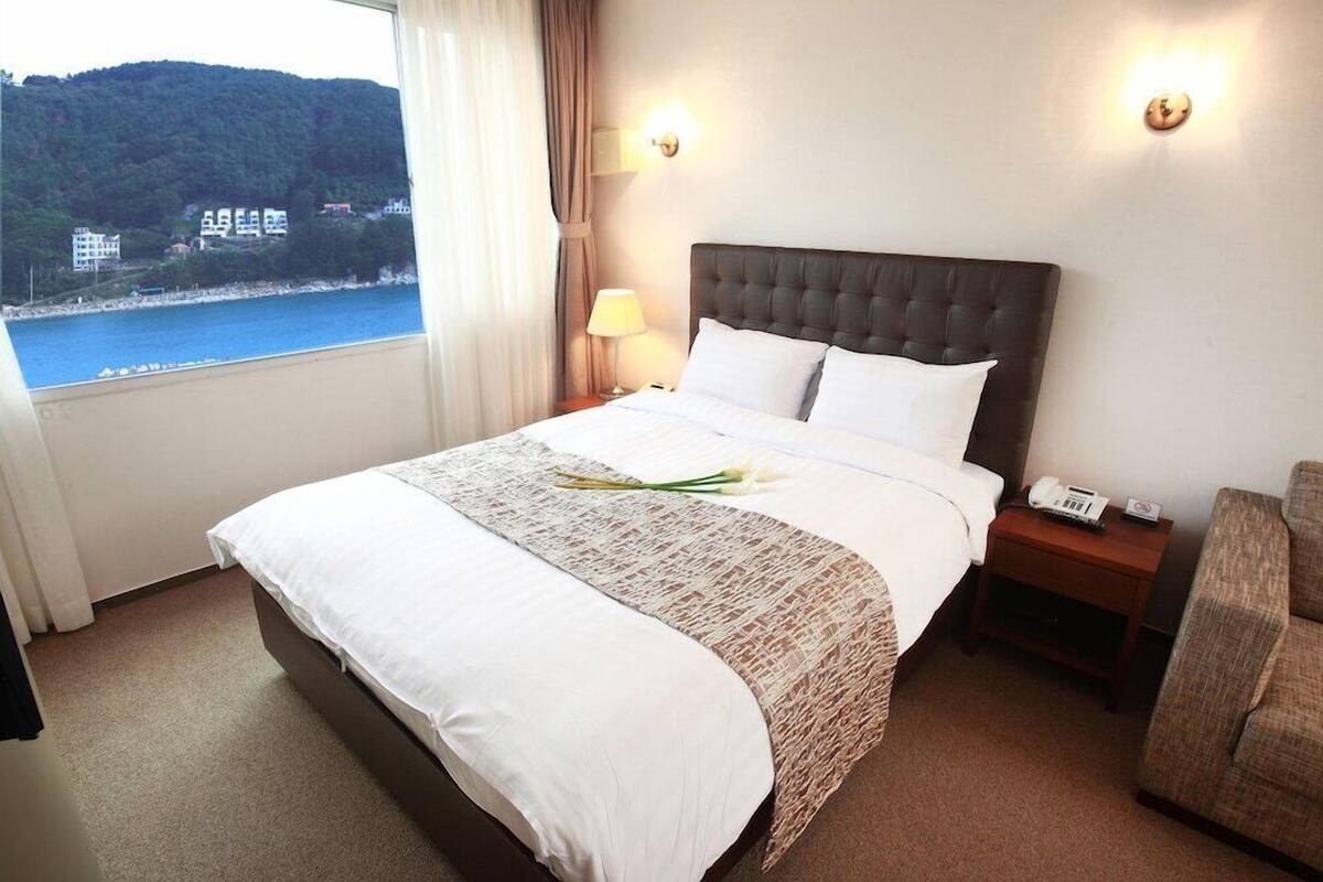 A hotel room with a bed, a nightstand with a phone, a chair, and a window with a view of a body of water and buildings.