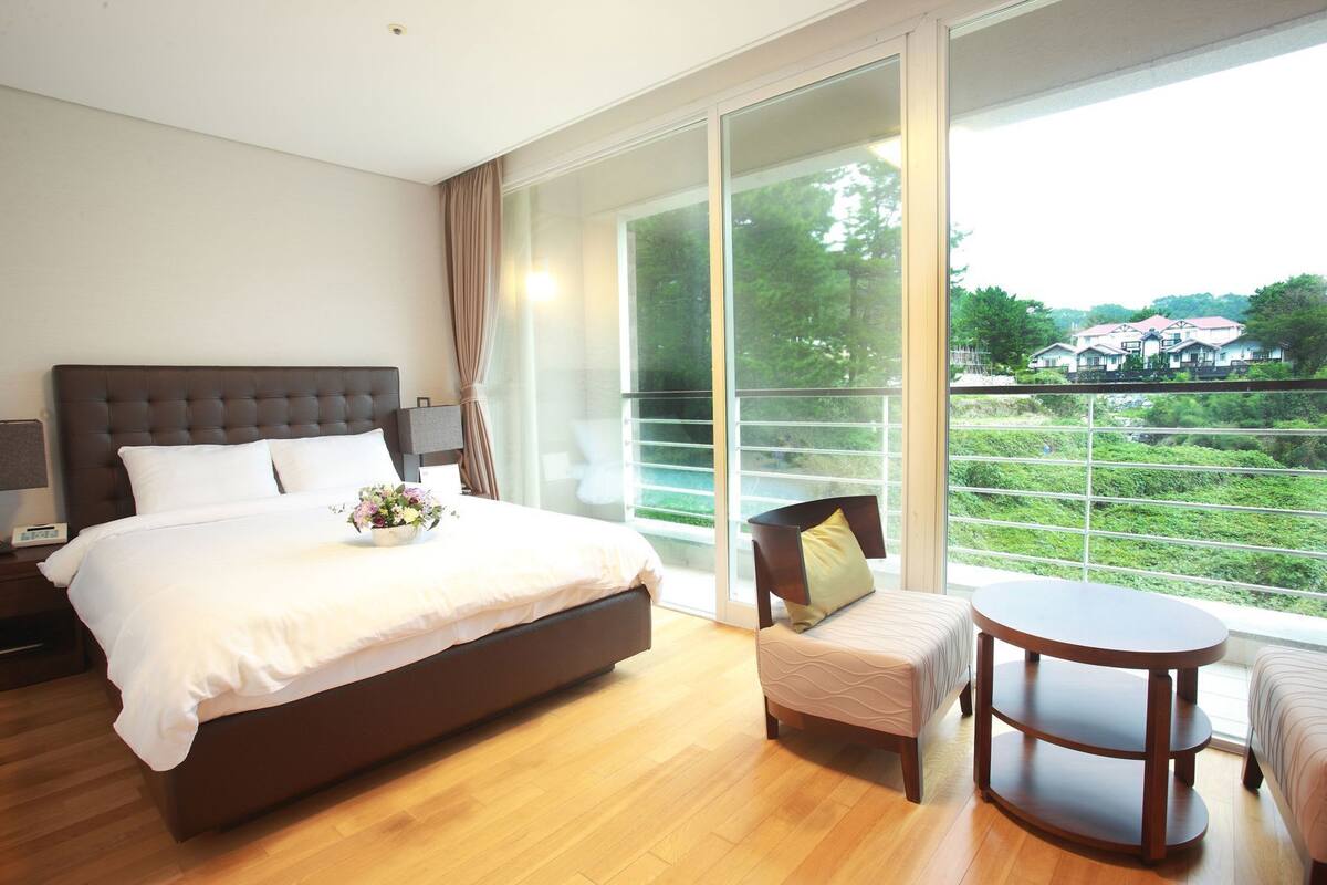 A modern hotel room with a large bed, a small sofa, a round table, and a balcony with a view of greenery and houses.