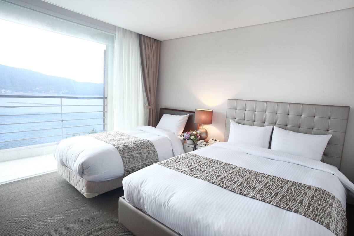 A hotel room with two beds, a large window with a view of the sea, and a nightstand with a lamp and flowers.