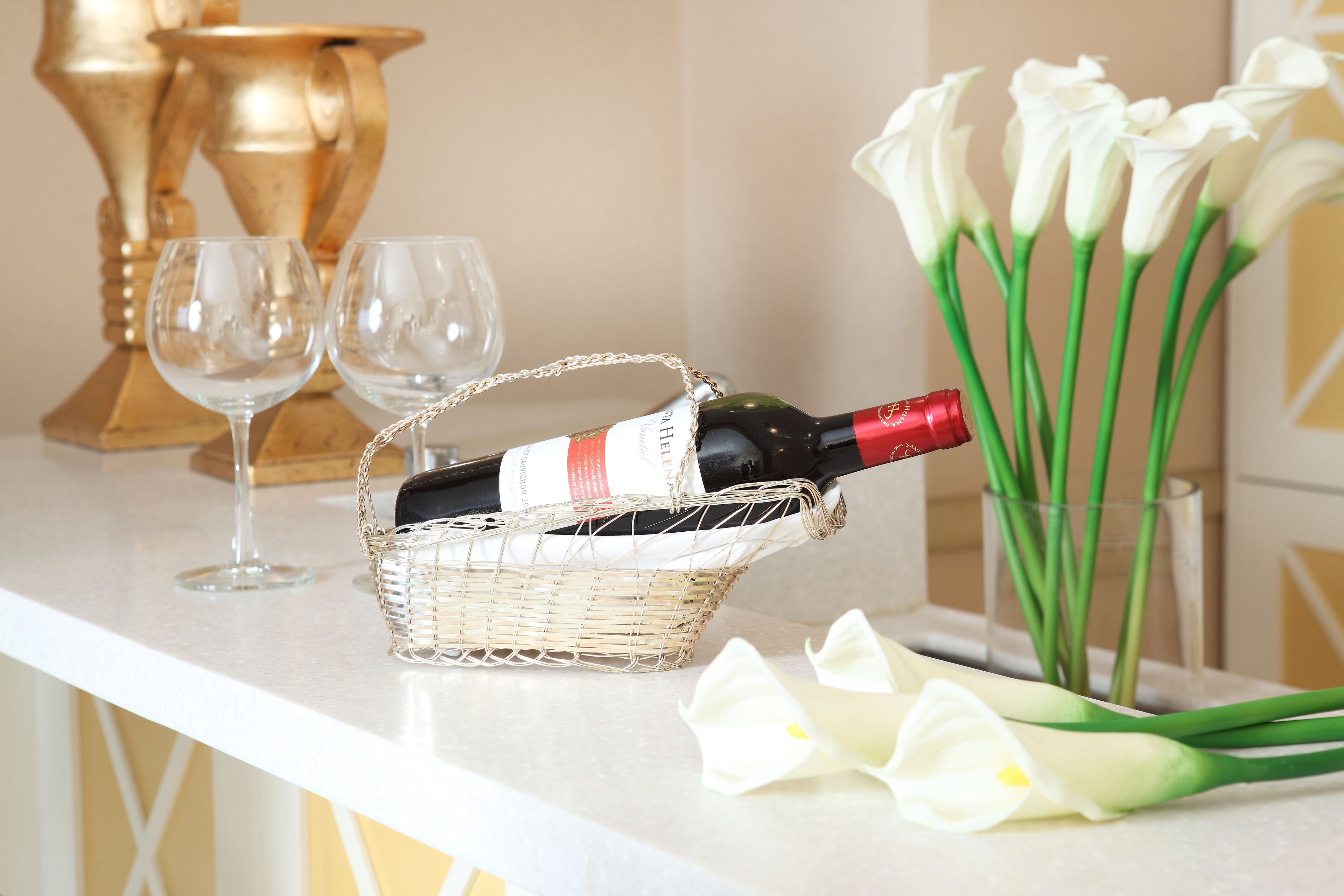 Una botella de vino en una cesta sobre una encimera, dos copas de vino vacías y un ramo de flores blancas.
