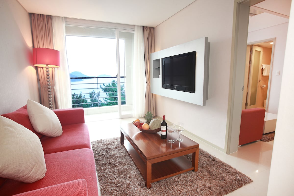 A modern hotel room with a red sofa, a wooden coffee table with a fruit bowl, a bottle of wine, and two glasses. A flat-screen TV is mounted on the wall, and a large window offers a view of the sea.