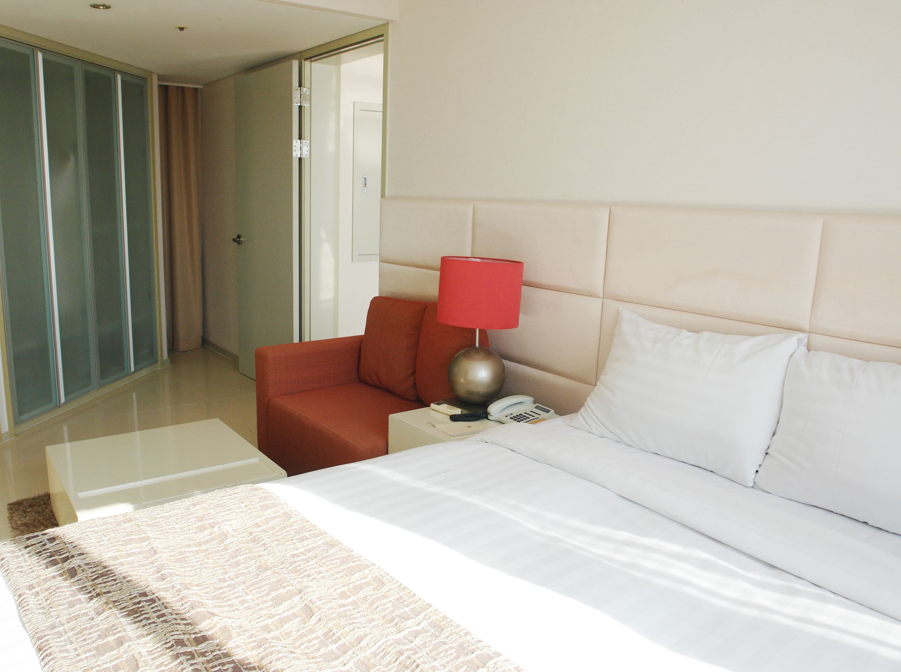 A hotel room with a bed, a red chair, a nightstand with a lamp, and a telephone.