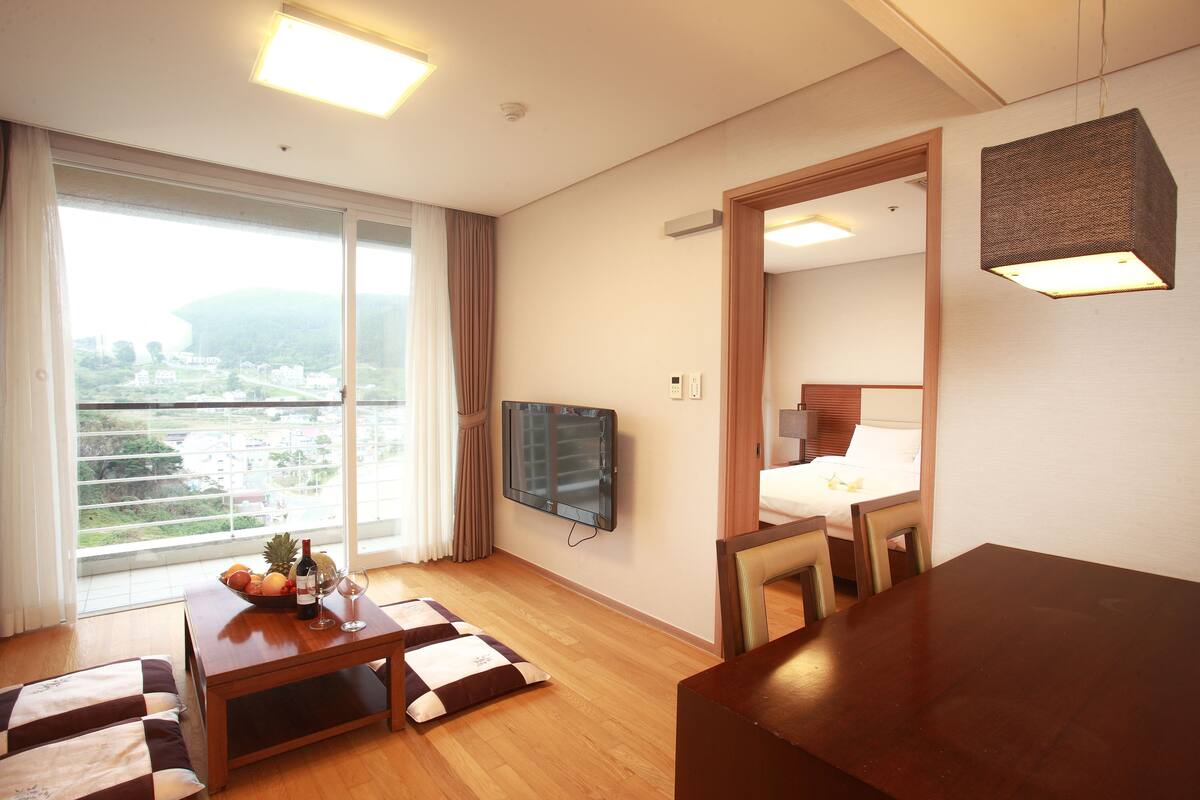 A hotel room with a large window, a flat-screen TV, a wooden dining table, and a bed with a wooden headboard.