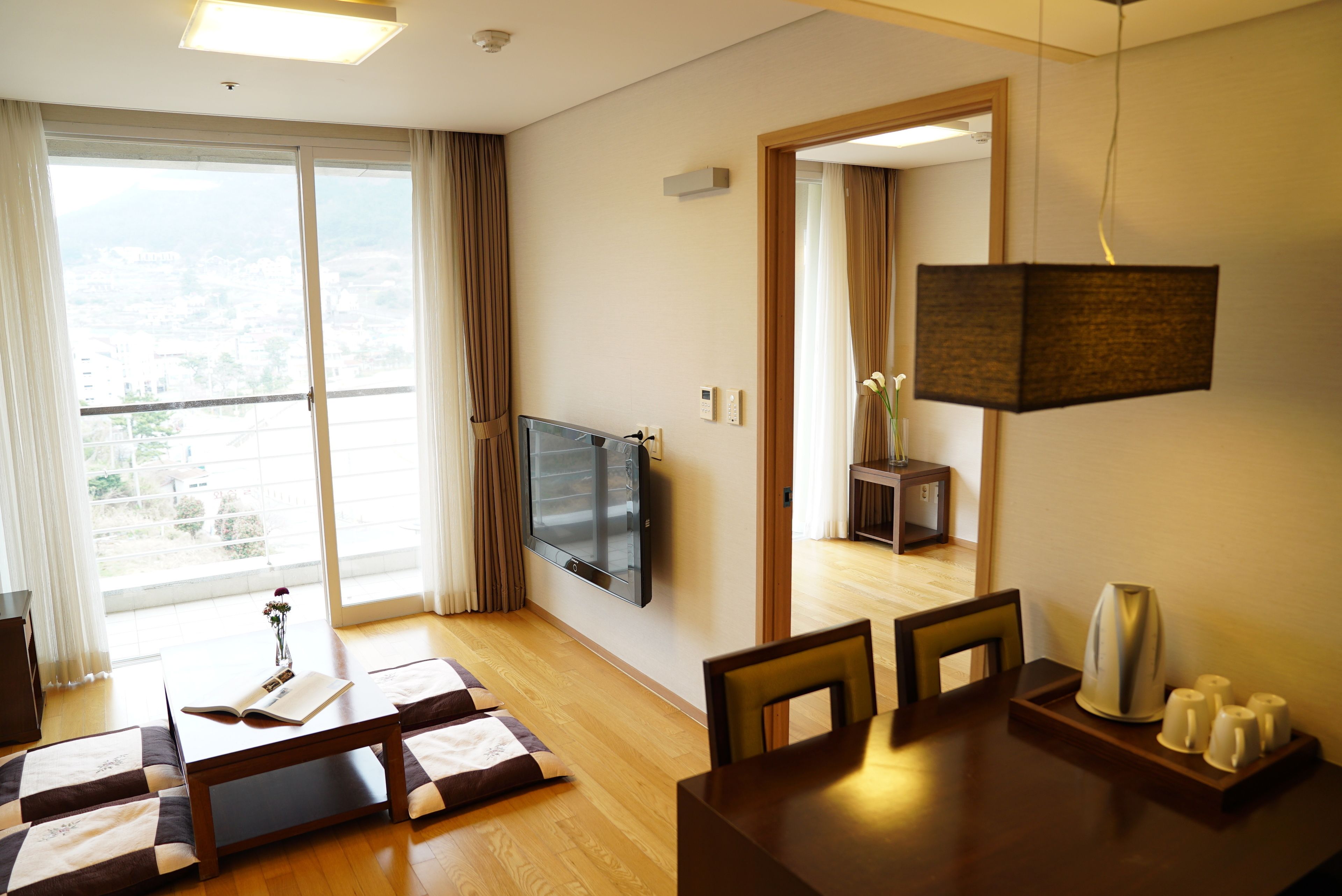 A modern living room with a flat-screen TV, a dining area with a wooden table and chairs, and a large sliding glass door leading to a balcony.