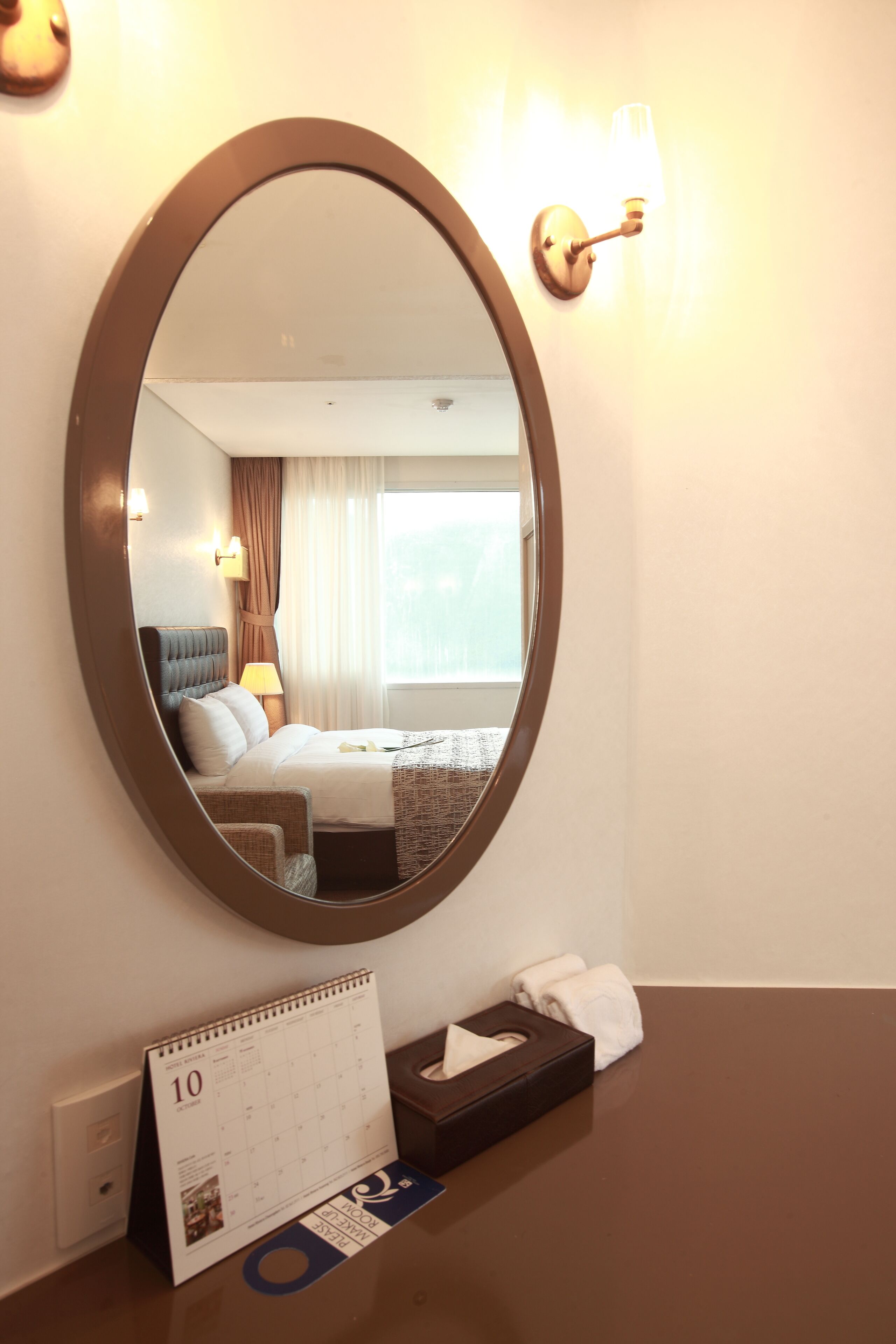 A hotel room with a large oval mirror, a bed with a headboard, a nightstand with a lamp, a calendar, and a tissue box.
