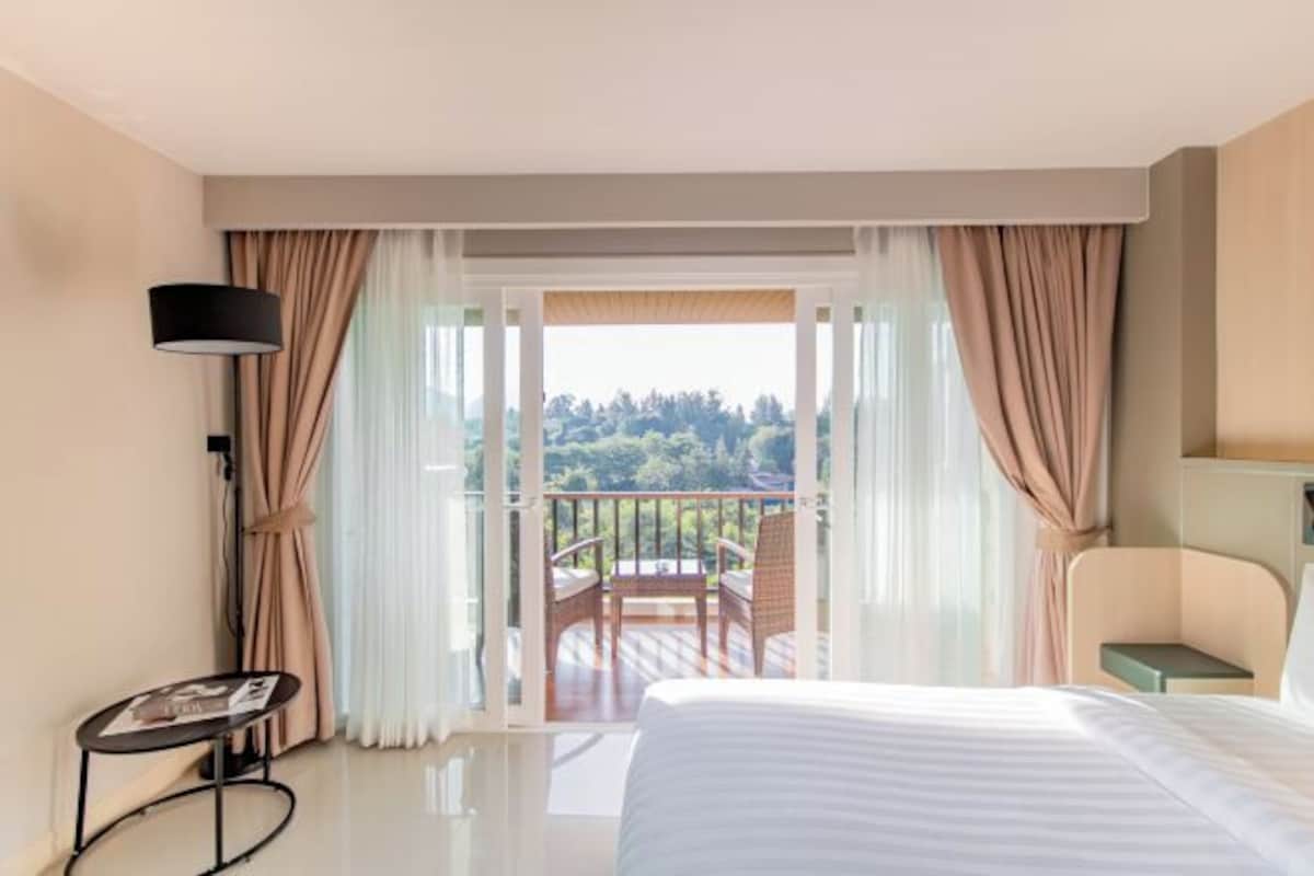 A hotel room with a bed, a small table, a chair, a lamp, and a balcony with a view of trees.