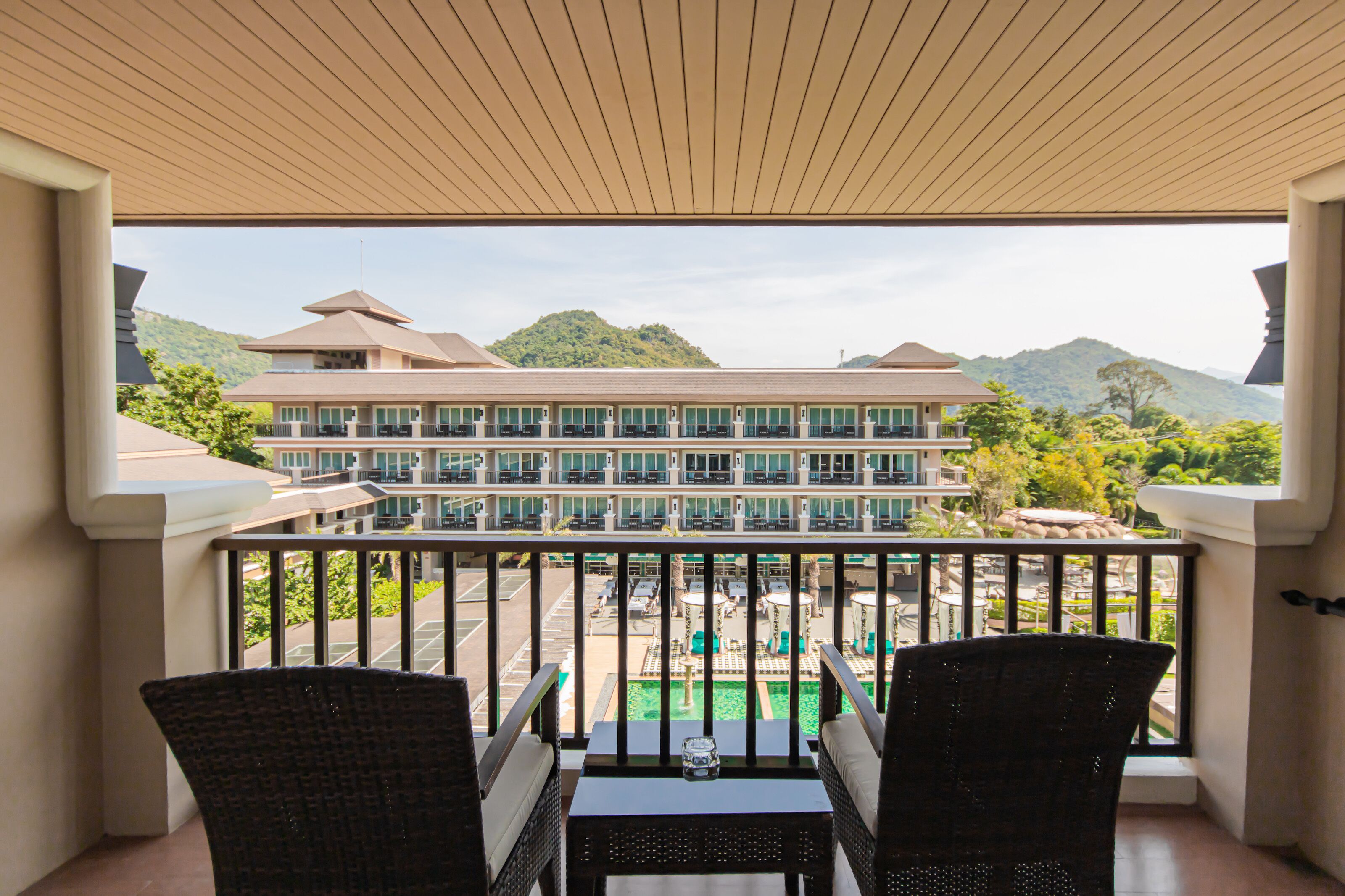 Un balcon agrémenté de chaises et d’une table en osier, d’où l’on peut admirer une piscine et un bâtiment d’hôtel à plusieurs étages, avec une chaîne de montagnes en toile de fond.