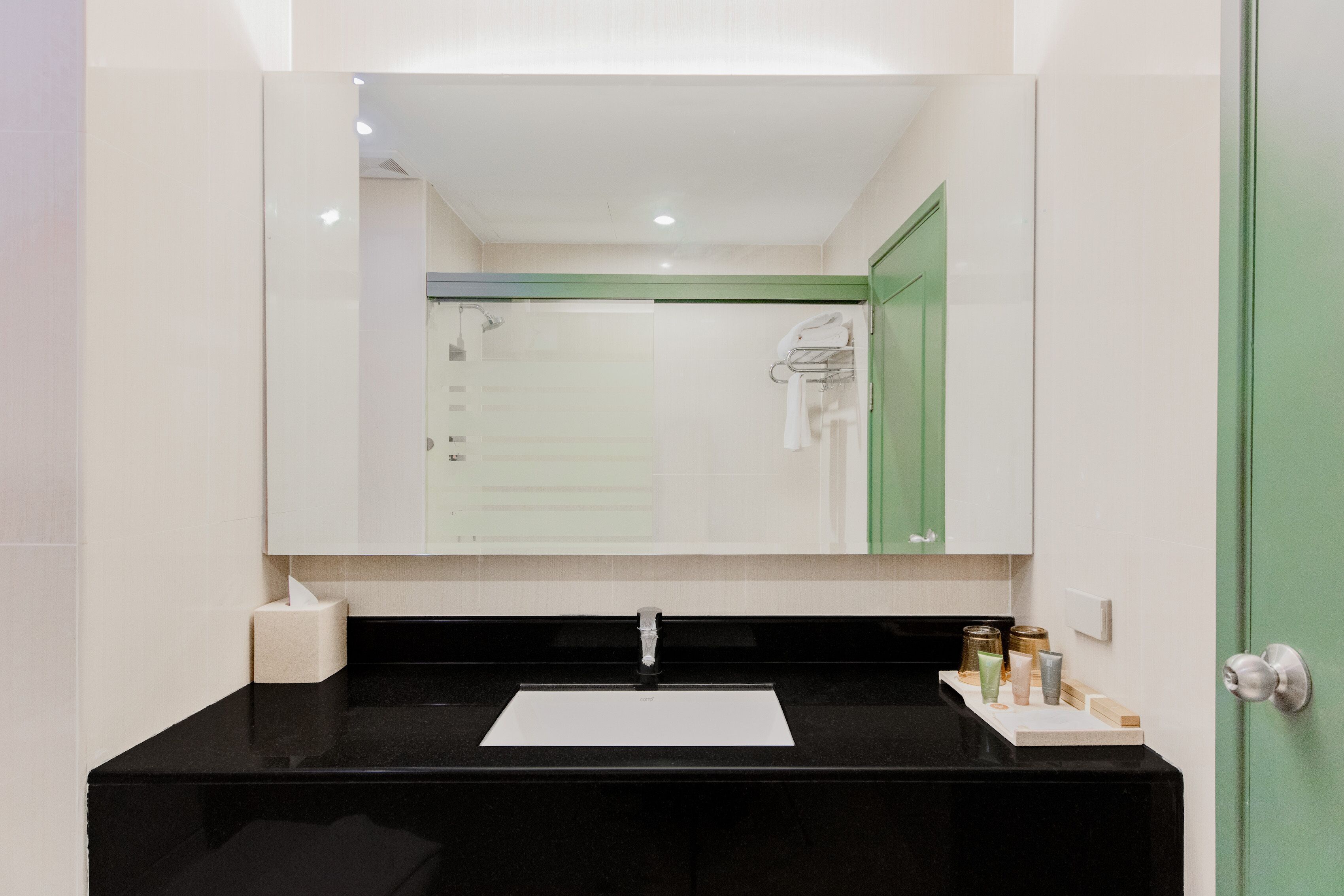 A bathroom with a black countertop, a white sink, and a mirror above it. There is a towel rack on the wall and a door with a green frame.