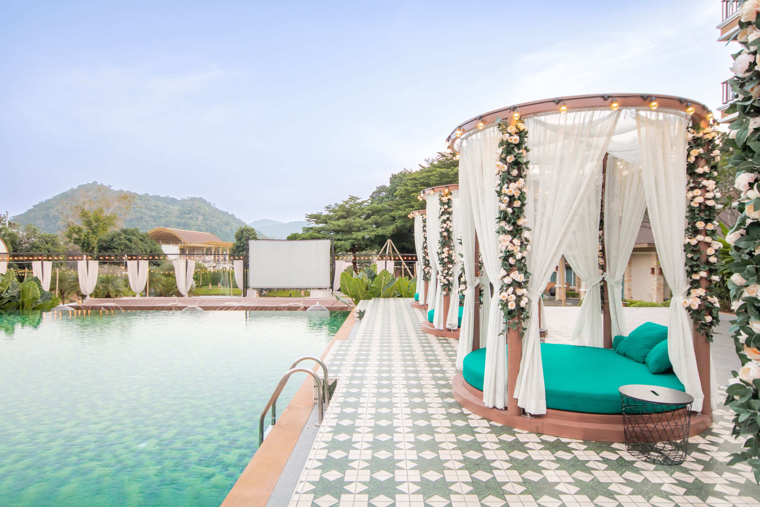A poolside area with a bed under a canopy, surrounded by greenery and a view of hills.