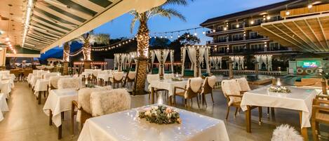 Un área de comedor al aire libre con mesas dispuestas para los invitados, palmeras y luces colgantes.