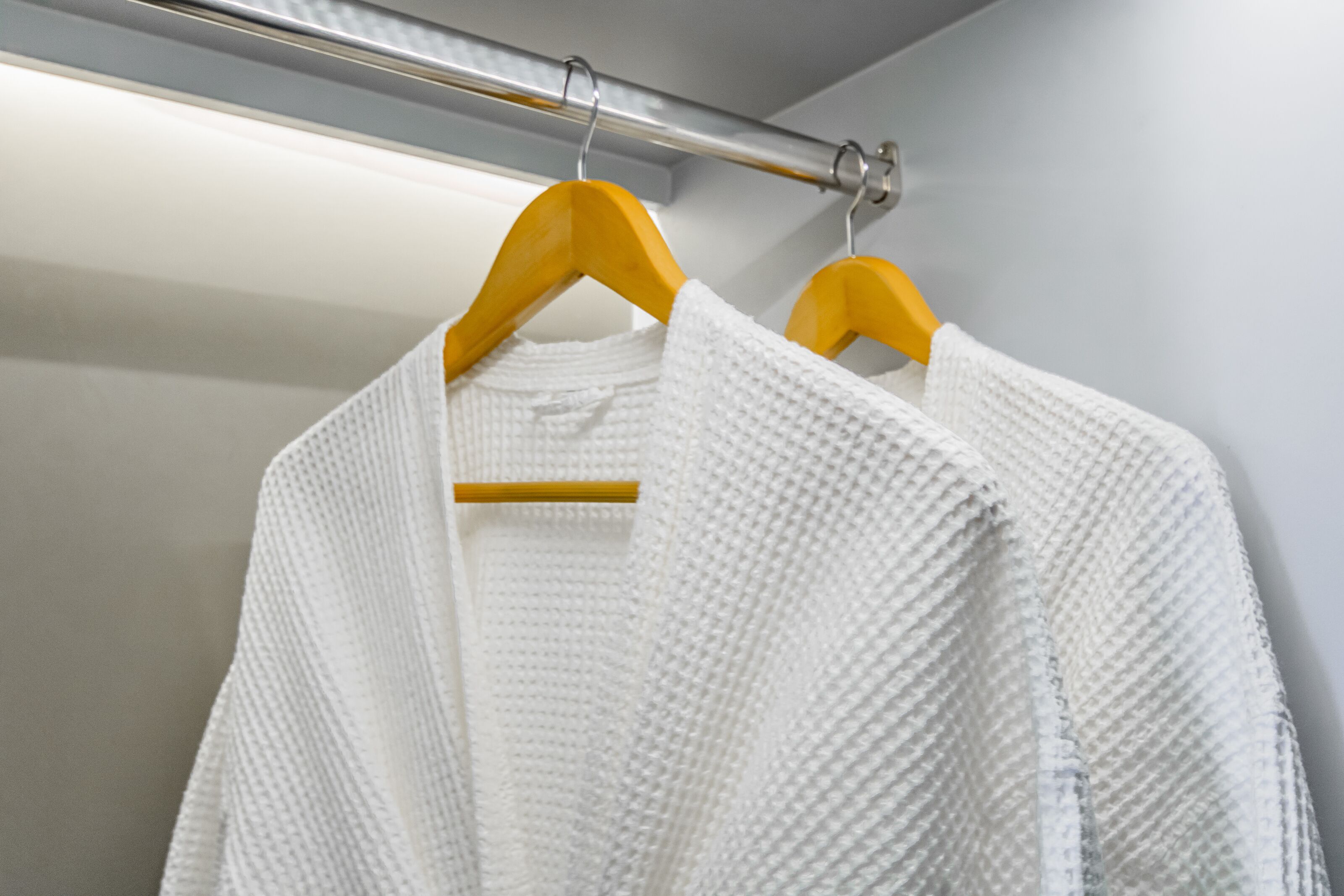 A white textured garment hanging on a wooden hanger in a room with a metallic rod and a white wall.