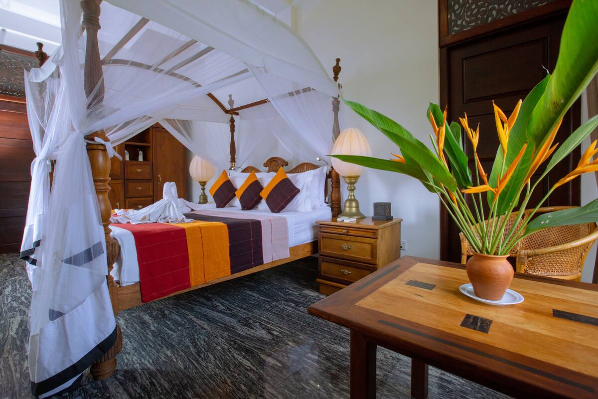 A room with a four-poster bed, a wooden nightstand, a table with a potted plant, and a wicker chair.