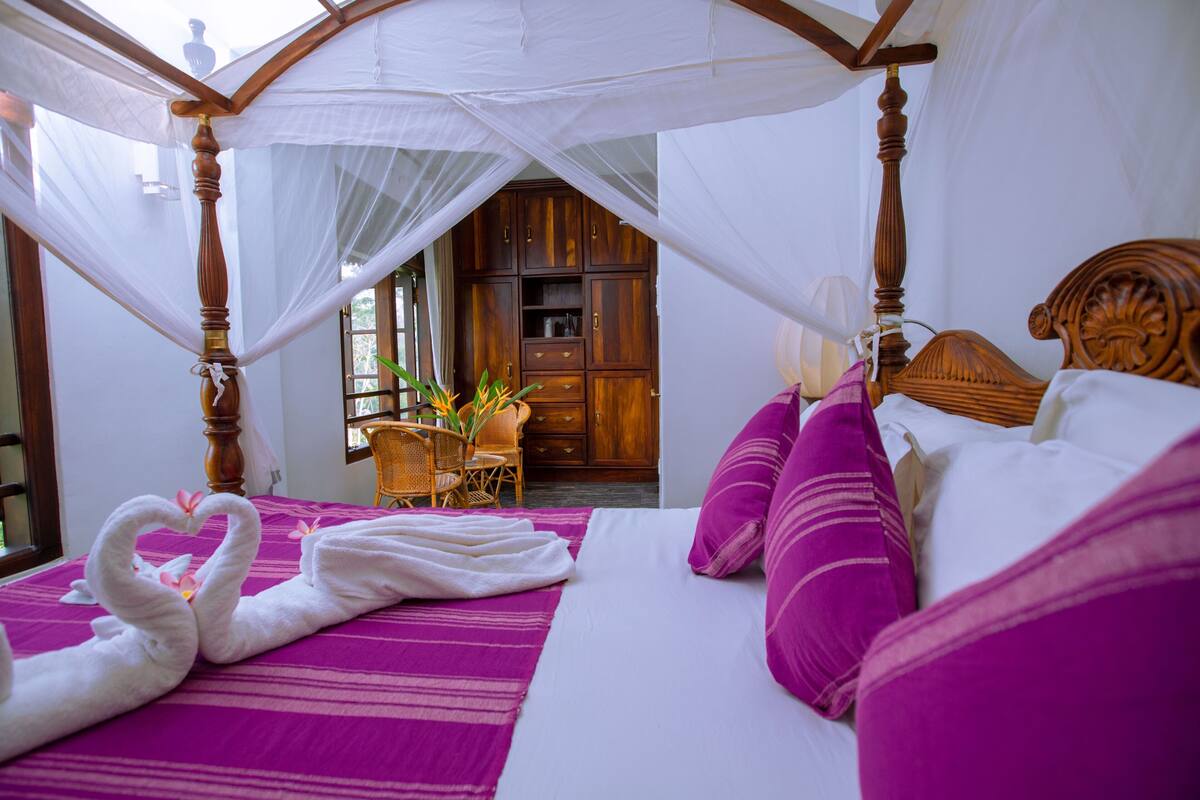 A room with a canopy bed, a wooden wardrobe, and a chair with a plant.