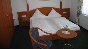 A hotel room with a double bed, a wooden nightstand, a chair, a small table with a plant, and a painting on the wall. - Hotel Garni Alt Refrath (Bergisch Gladbach)