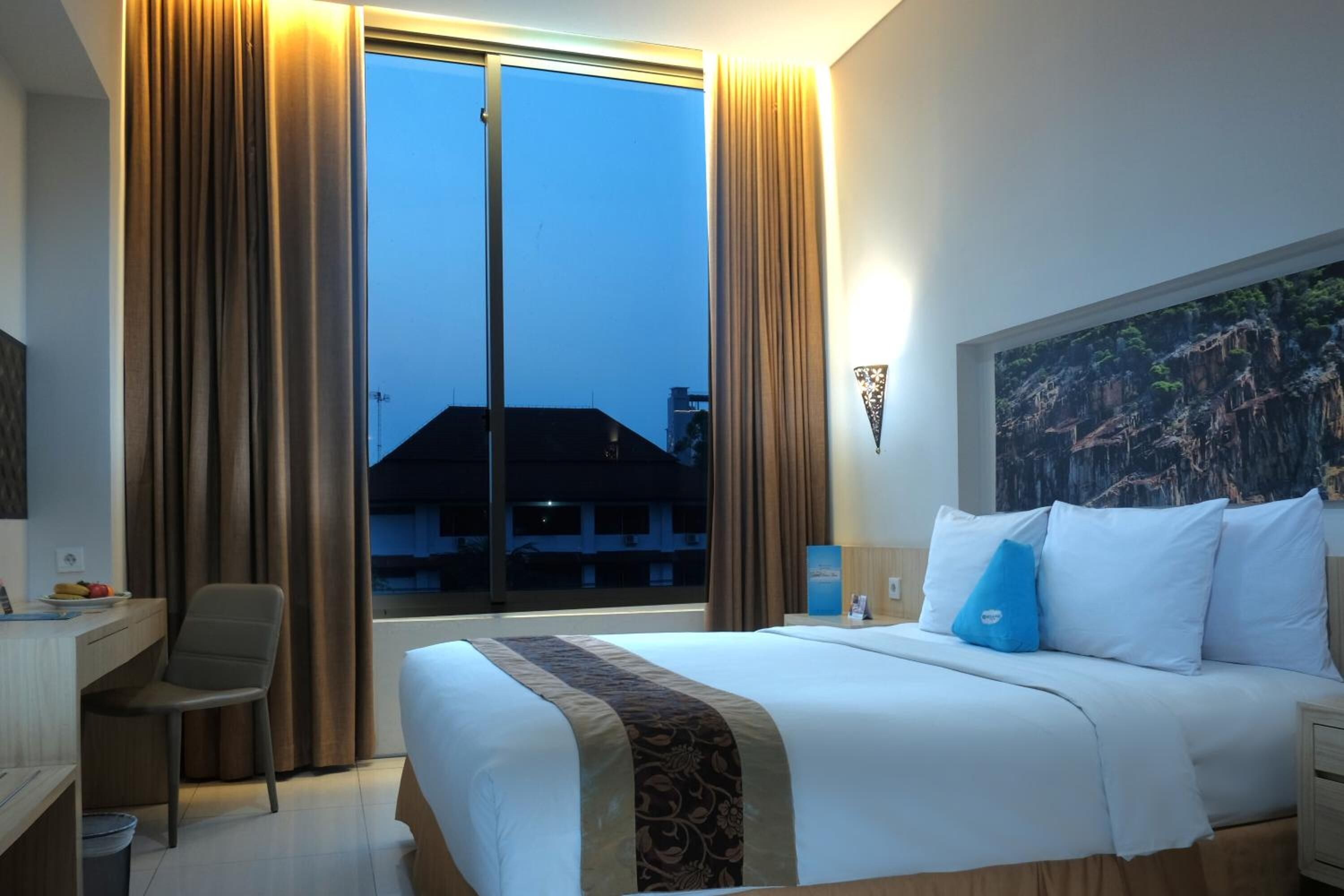 A hotel room with a large bed, a desk, and a chair. There is a window with curtains, a painting on the wall, and a view of buildings outside.