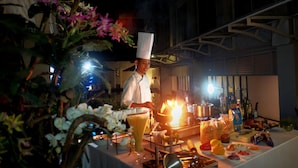 A chef cooking outdoors at night with a table full of ingredients and a decorative plant in the foreground. - Megaland Hotel (Surakarta)