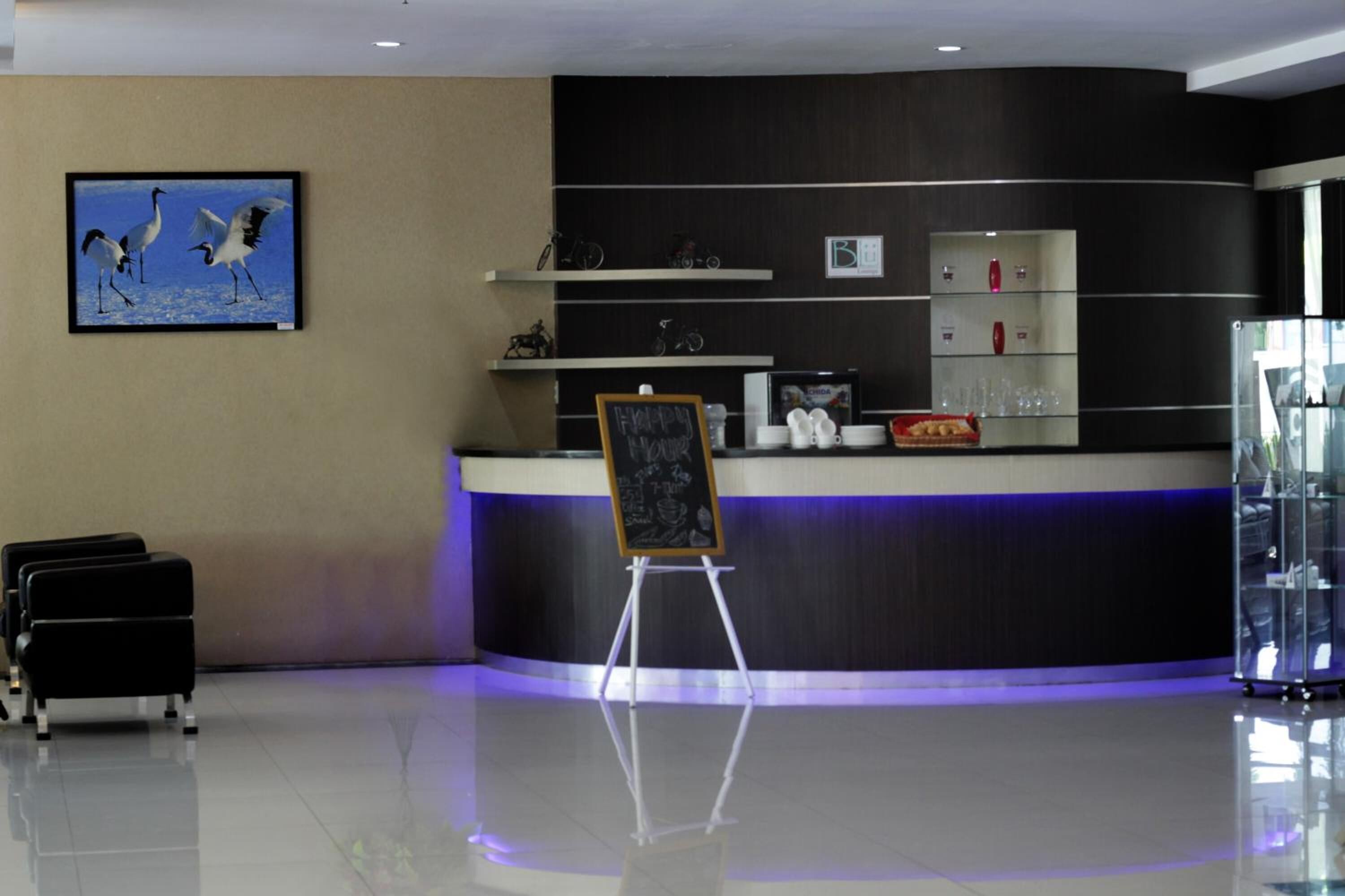 Zona de recepción de hotel con mostrador, una pizarra y un cuadro enmarcado con pájaros. Se observan estantes con objetos y una vitrina de vidrio.