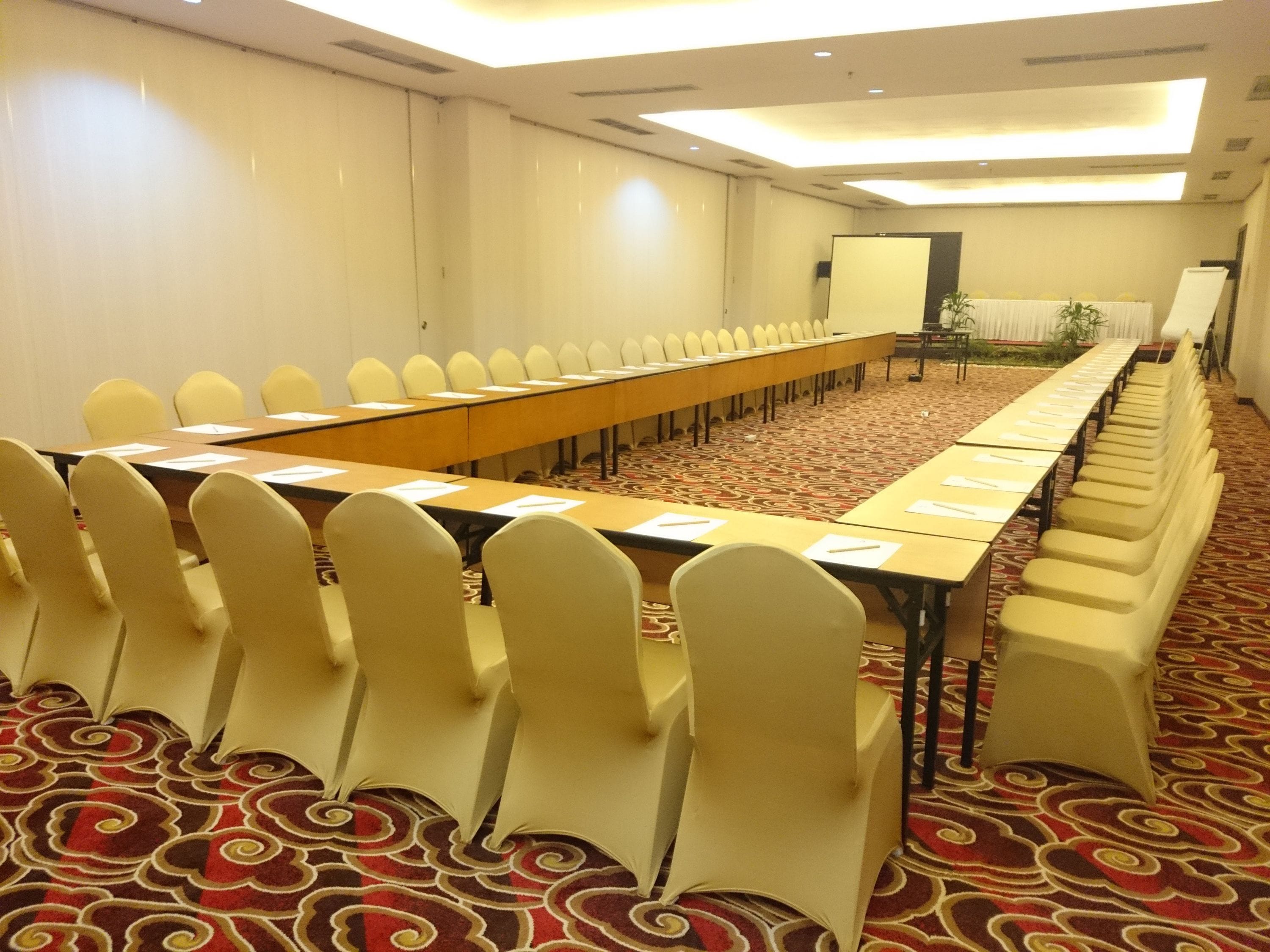 Sala de conferências com uma mesa longa e cadeiras, piso coberto de carpete e tela de projetor.
