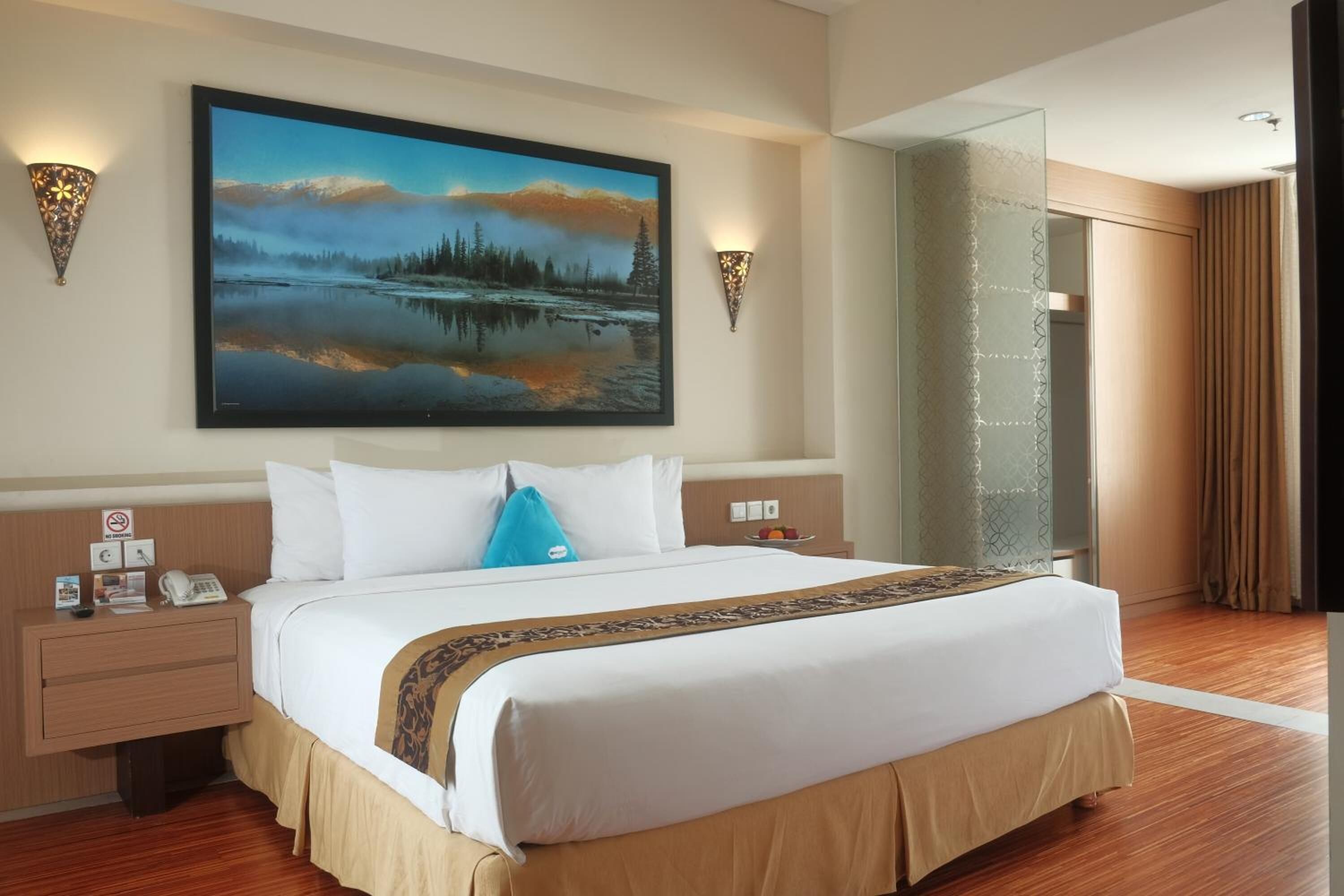 Una habitación de hotel con una cama grande, dos mesitas de noche, un buró, un televisor montado en la pared y un cuadro de un paisaje lacustre.