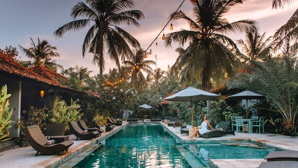 A pool area with lounge chairs and umbrellas, surrounded by palm trees and a tiled roof. - Belukar Villas (Gili Trawangan)
