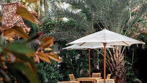 A poolside area with lounge chairs and a table under a large umbrella, surrounded by lush tropical plants and palm trees. - Belukar Villas (Gili Trawangan)