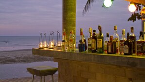 A bar with bottles and glasses on a wooden deck overlooking the beach at dusk.