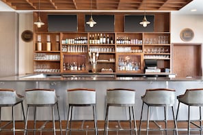 A bar area with a long counter, multiple shelves stocked with bottles and glasses, and three bar stools. - Four Points by Sheraton Fargo Medical Center (Fargo)