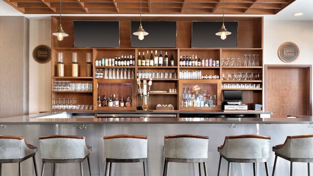 A bar area with a long counter, multiple shelves of bottles and glasses, and four bar stools. - Four Points by Sheraton Fargo Medical Center (Fargo)