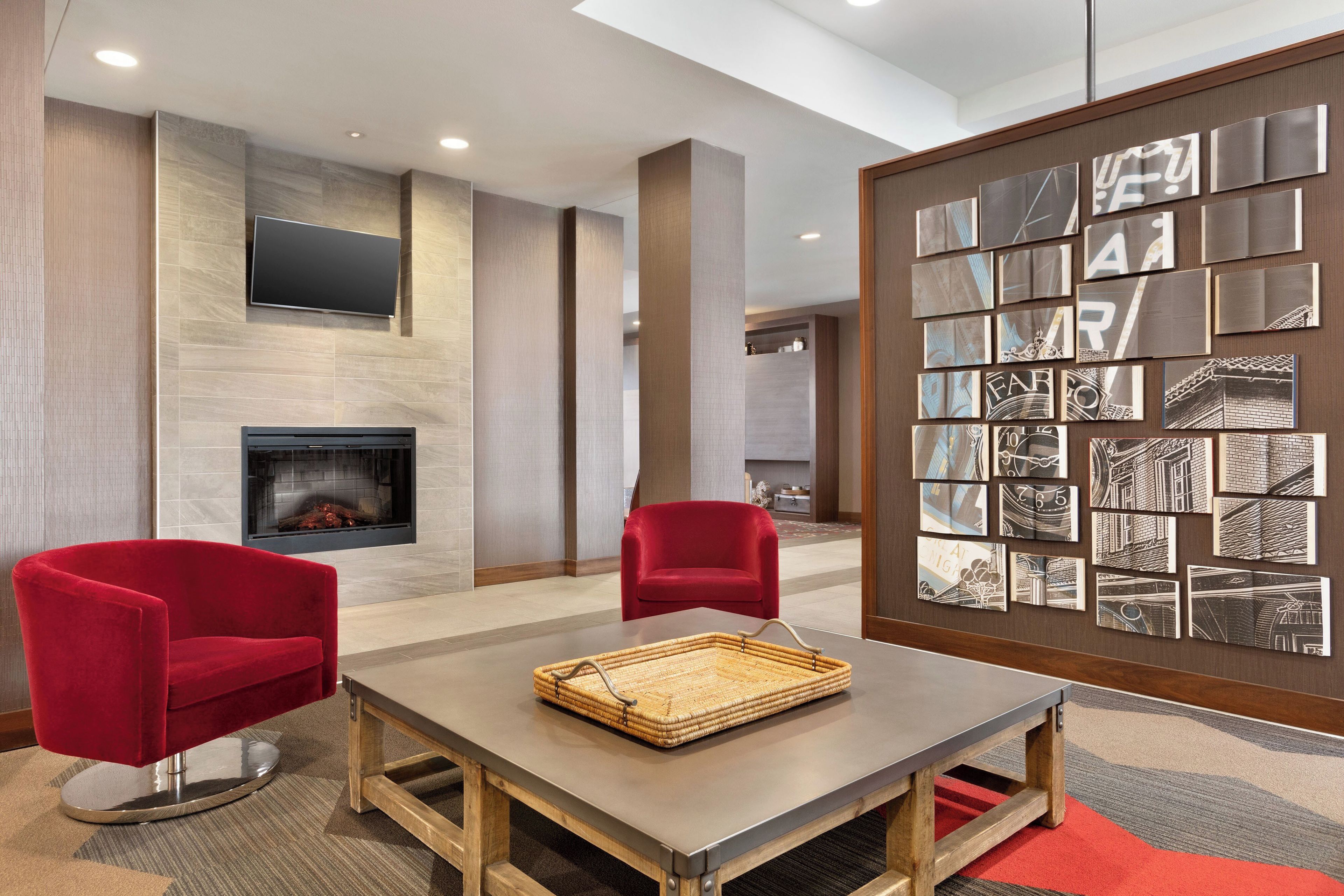 A modern lobby with a fireplace, red chairs, a table with a woven tray, and a wall with framed artwork.