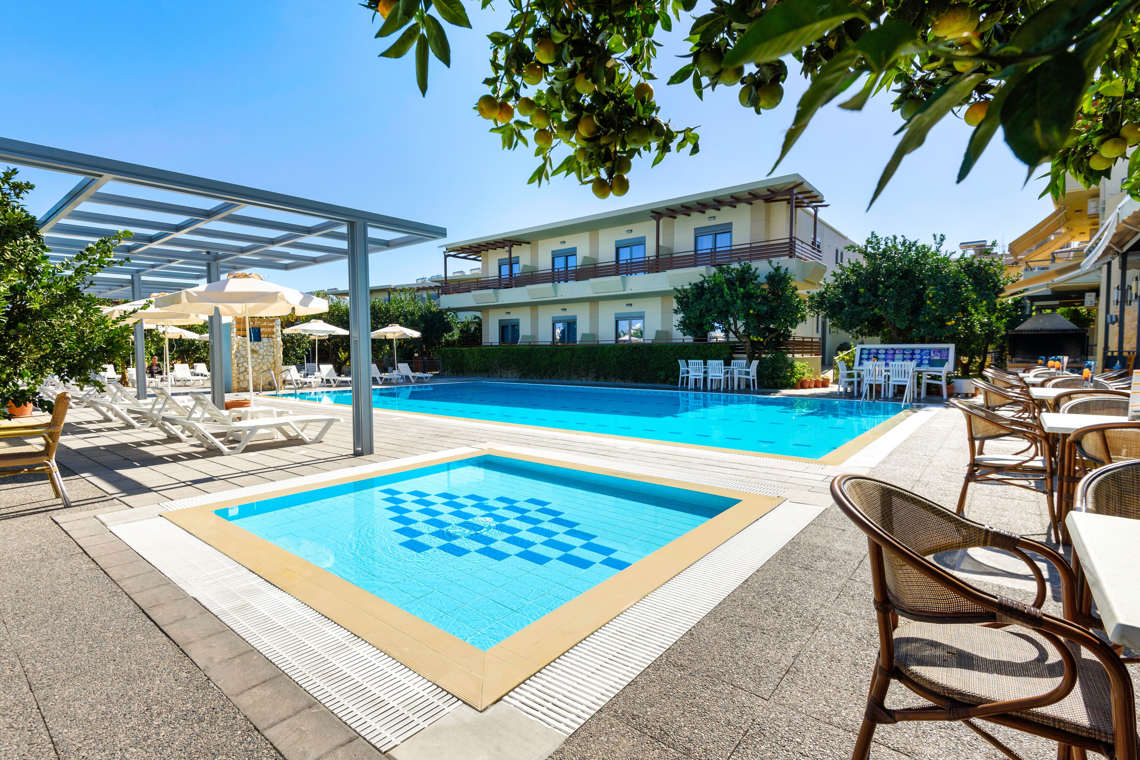 Uma área de piscina com espreguiçadeiras e guarda-sóis, um deck de piscina xadrez e um prédio com varandas ao fundo.