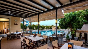 Un área de comedor al aire libre con mesas y sillas, una piscina y vistas a un edificio azul en el fondo.