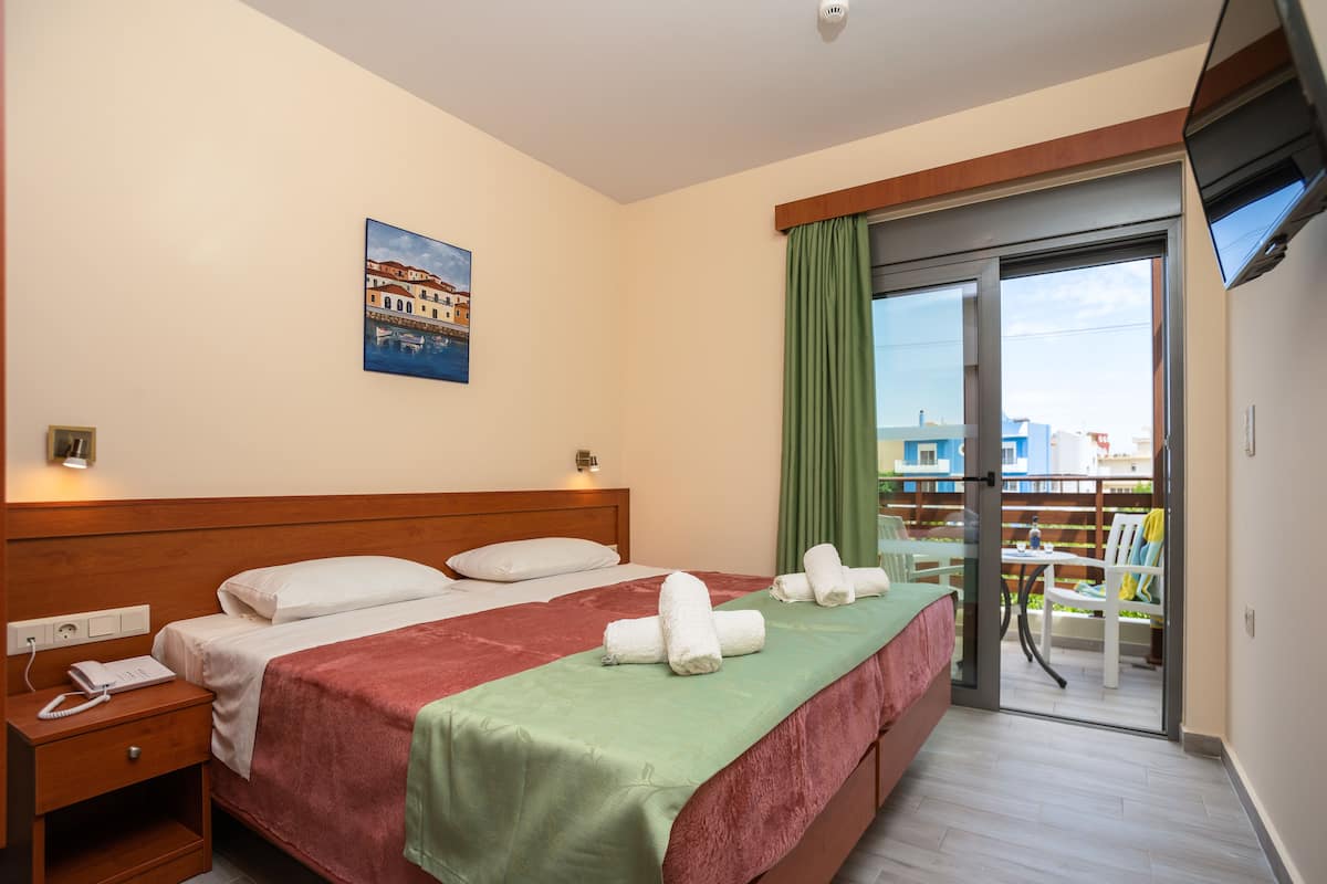 A hotel room with a bed, a nightstand, a telephone, a painting on the wall, and a balcony with a view of buildings.