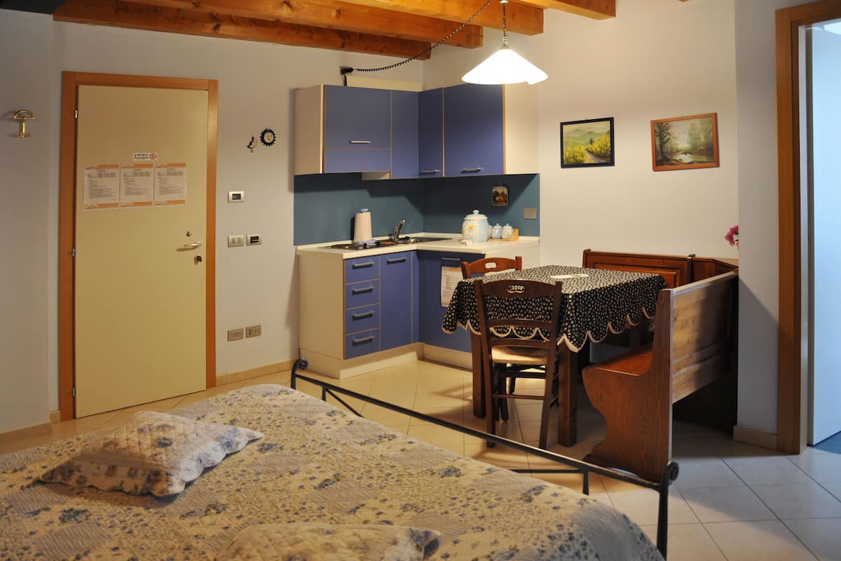 A bedroom with a bed, a wooden chair, a small table with a tablecloth, a kitchen area with cabinets, and a door with a sign.
