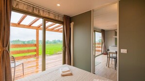 A bedroom with a large window, wooden ceiling beams, a bed with towels, and a view of a green field.