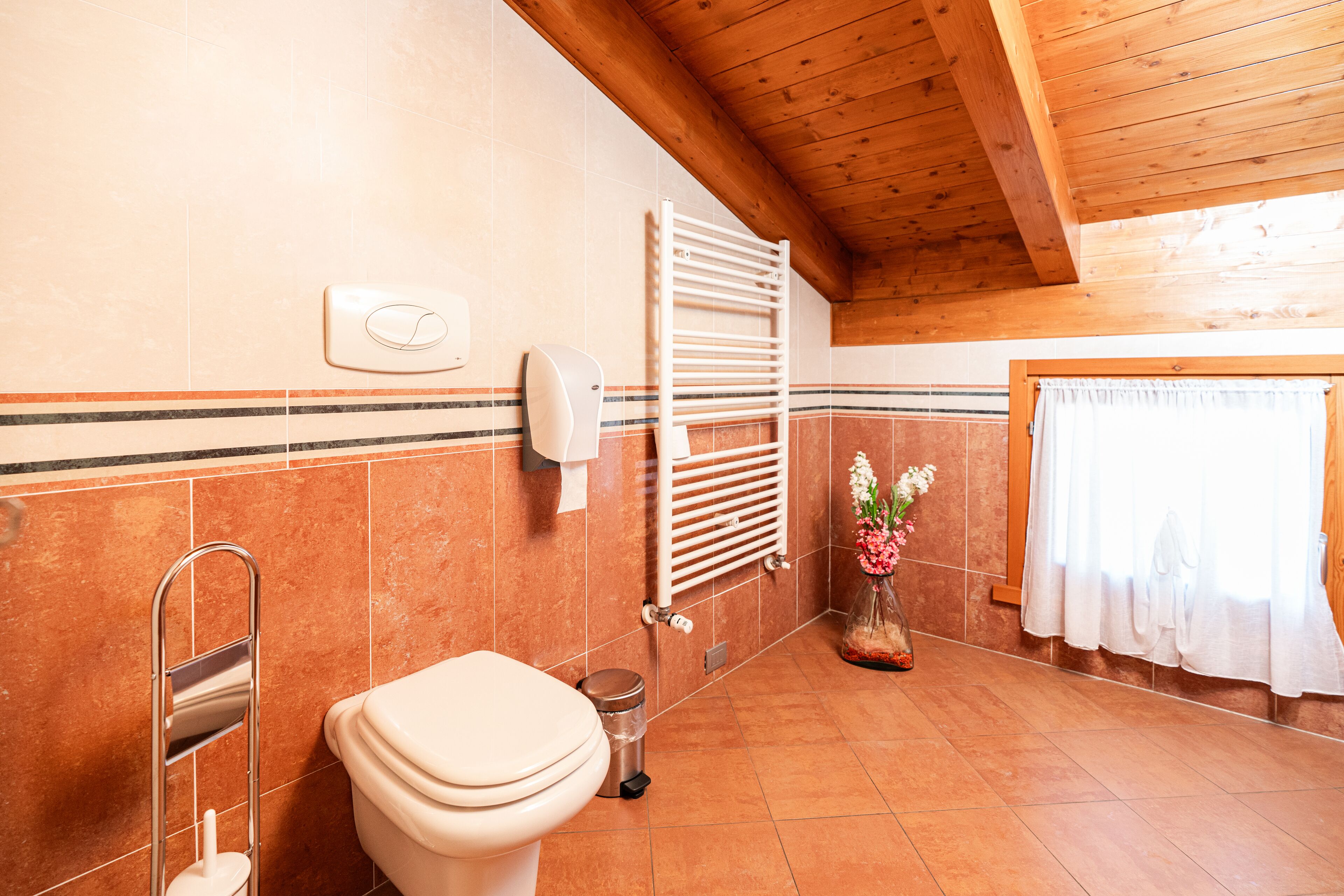 A bathroom with a toilet, a hand dryer, a towel rack, a waste bin, and a window with a curtain.