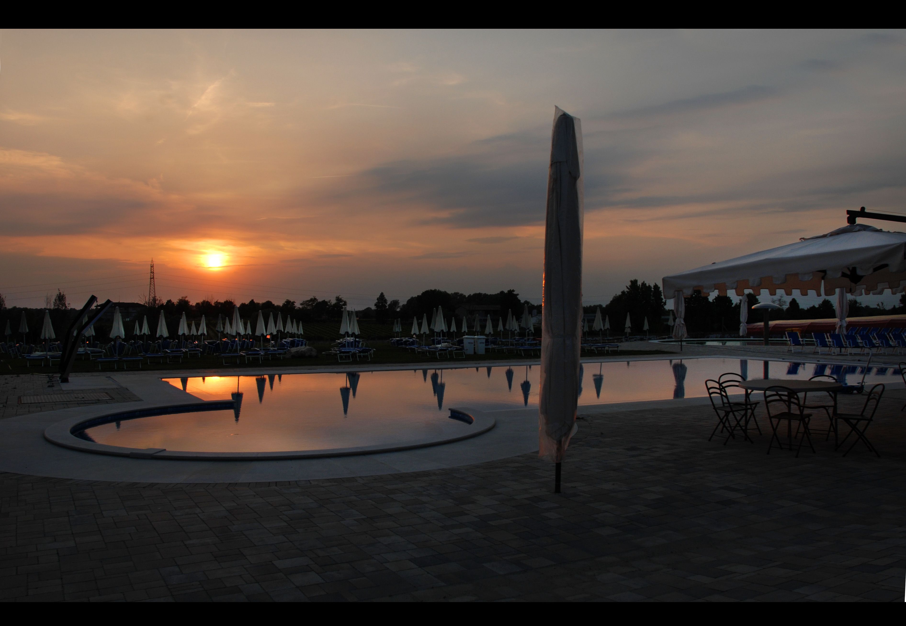 Atardecer sobre una piscina con sillas de descanso y sombrillas, una escultura alta y un pabellón.