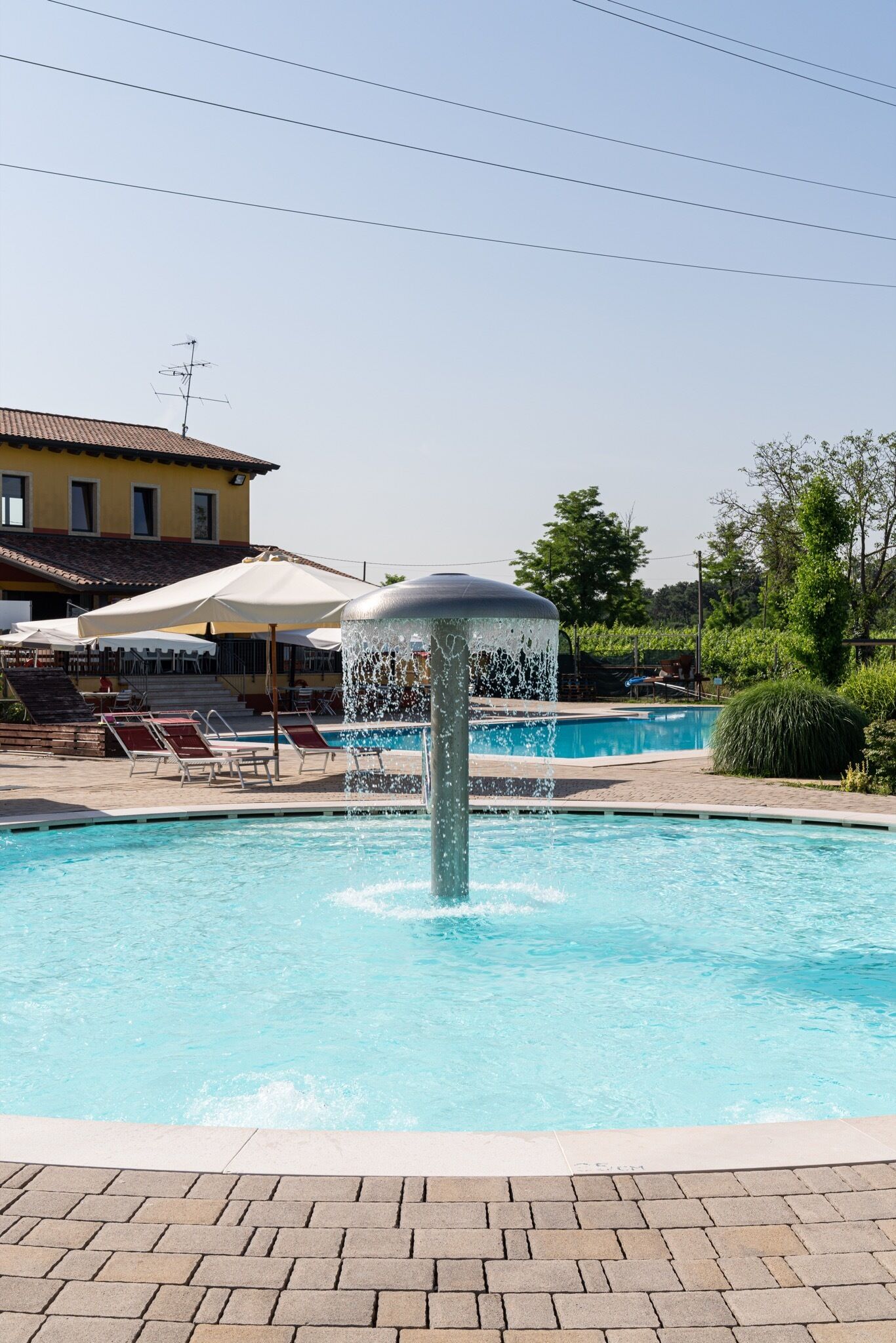 Une piscine avec une fontaine, des chaises longues et, au fond, un bâtiment doté d’un toit recouvert de tuiles.
