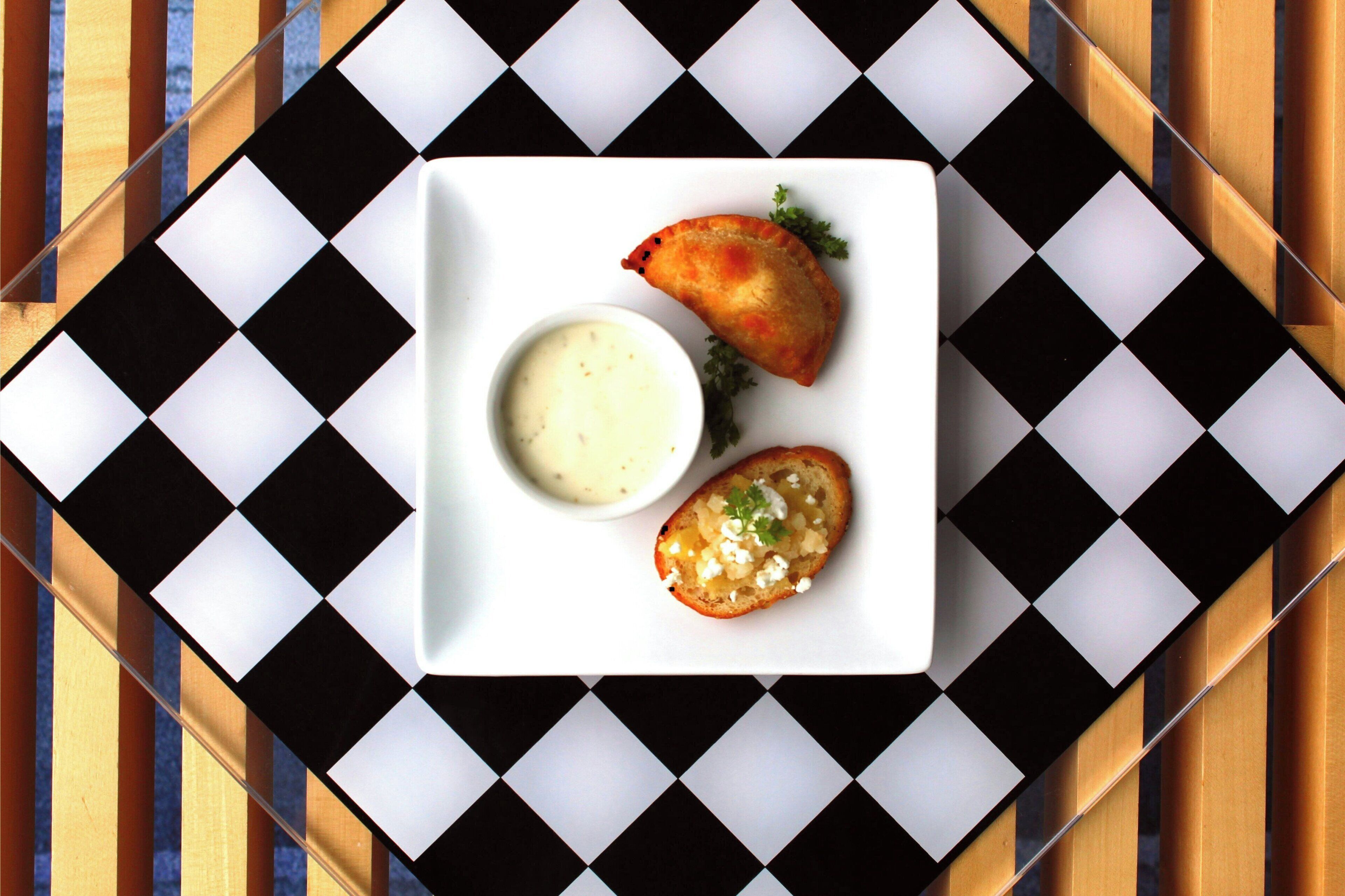 A white plate with two pastries and a small bowl of sauce on a checkered tablecloth.