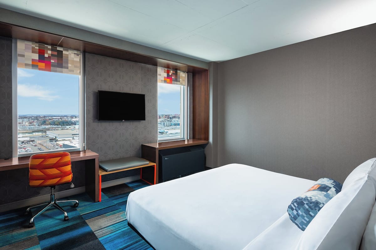 A hotel room with a bed, a desk with a chair, a television, and a window with a city view.