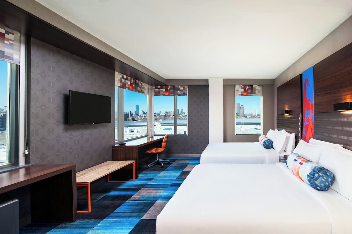 A hotel room with a city view, a desk with a chair, two beds, and a flat-screen TV mounted on the wall.