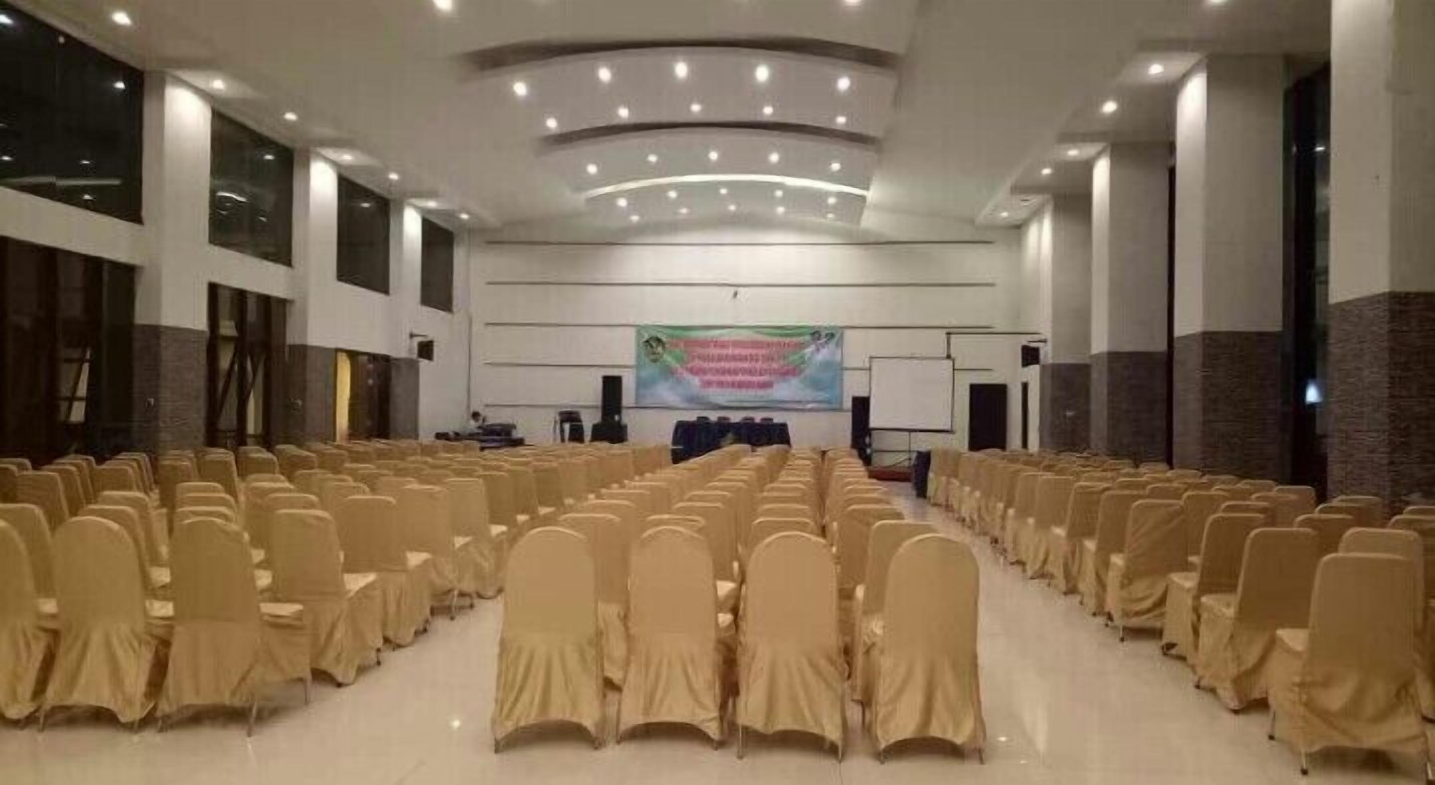 Un salón amplio con filas de sillas tapizadas en tono beige, un escenario con un cartel y una pizarra.
