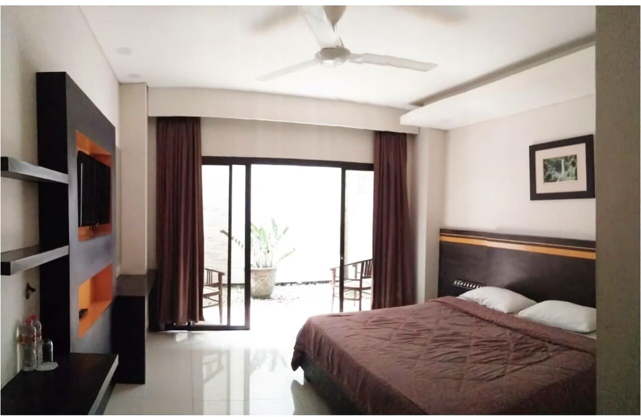 Habitación de hotel con cama, ventilador de techo, televisor, puerta corrediza a un balcón y un cuadro en la pared.