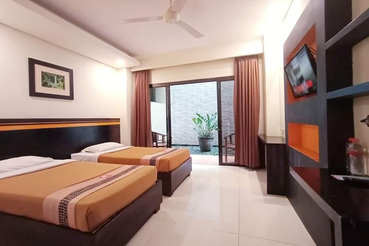 Une chambre d’hôtel avec deux lits, un ventilateur de plafond, une grande fenêtre avec des rideaux, une plante en pot et un téléviseur posé sur un meuble.