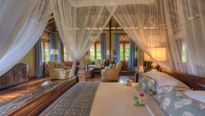 A spacious bedroom with a large bed, a sitting area with a sofa and chairs, and a view of the outdoors through large windows. - Kahanda Kanda (Angulugaha)