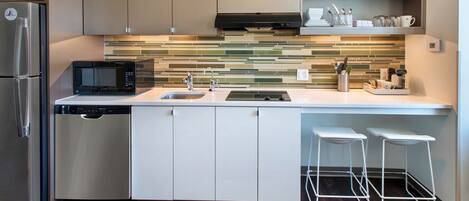 A modern kitchen with a stainless steel refrigerator, a dishwasher, a microwave, a sink, and a stove. There are two white bar stools and a tiled backsplash.
