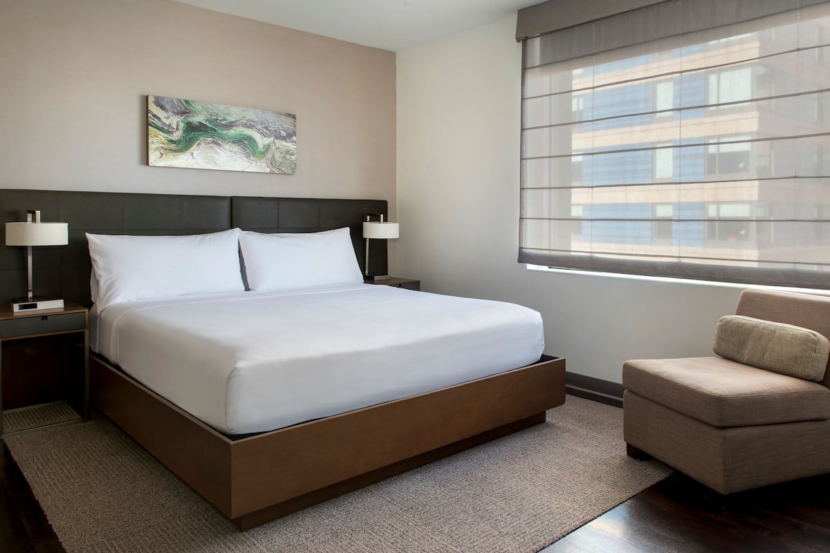 Une chambre d’hôtel moderne, dotée d’un grand lit, de deux tables de chevet avec des lampes, d’un petit fauteuil et d’une fenêtre avec des stores.