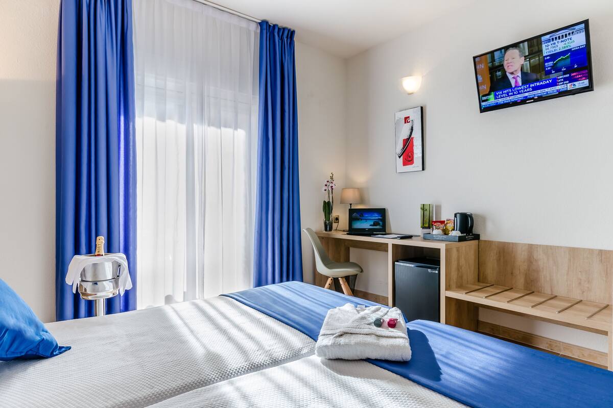 A hotel room with a bed, a desk with a computer, a chair, a small refrigerator, and a TV on the wall.