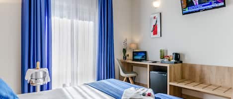 Ein Hotelzimmer mit einem Bett, einem Schreibtisch mit Computer, einem Sessel, einem kleinen Kühlschrank und einem Fernseher an der Wand.
