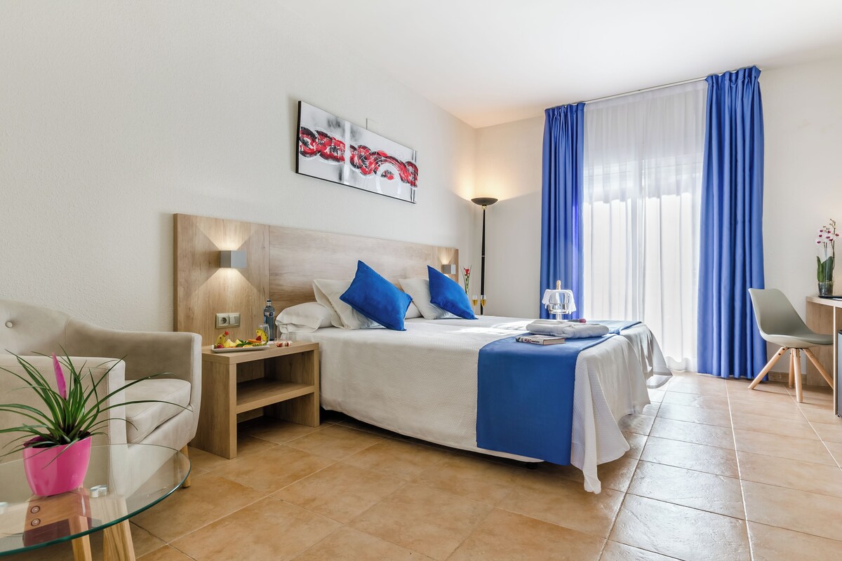 A modern hotel room with a bed, a sofa, a glass table, and a potted plant.